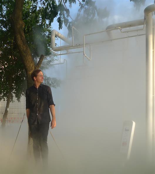 Philippe Rahm in a cloud of water vapour in the Meteorological Garden at the 11th Architecture Biennale in Venice. Pipes and trees emerge from the cloud in the background.